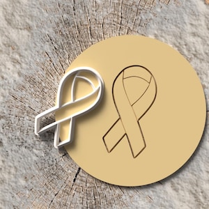 May include: A light yellow ribbon cookie cutter and a ribbon imprint on a round, light yellow surface. The objects are set against a textured, light brown and gray background.