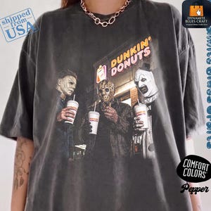 May include: A charcoal grey short-sleeve t-shirt featuring a graphic print of three horror movie characters standing in front of a Dunkin' Donuts store, each holding a white coffee cup. The shirt has a crew neckline and a relaxed, boxy fit.