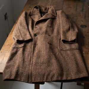 Puede incluir: Un abrigo de tweed marrón con diseño de espiga y doble botonadura. El abrigo presenta un cuello con solapas, dos bolsillos delanteros y cierres de botones. El abrigo mide aproximadamente 102 cm de largo.