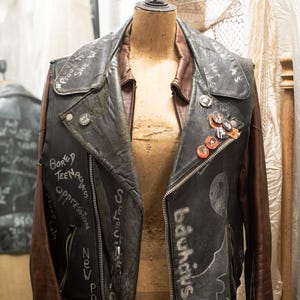 Puede incluir: Una chaqueta de cuero negra vintage con mangas y cuello marrones, exhibida en un maniquí. La chaqueta presenta texto escrito a mano y varios alfileres. El texto incluye palabras como "Bored Teenagers" y "Bauhaus".