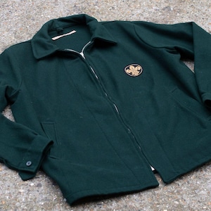 Puede incluir: Chaqueta verde oscuro con cremallera, cuello y un pequeño emblema en el pecho. La chaqueta tiene dos bolsillos delanteros y puños abotonados. La chaqueta está hecha de un material grueso, posiblemente lana.