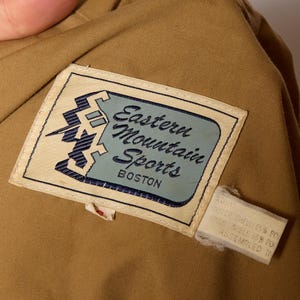 Op de afbeelding: Een beige stoffen jas met een bruin label waarop "Eastern Mountain Sports Boston" staat. Het label heeft een blauwe en witte afbeelding van een berg met een bliksem.