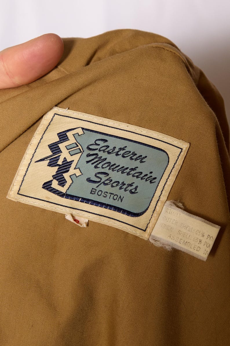 Op de afbeelding: Een beige stoffen jas met een bruin label waarop "Eastern Mountain Sports Boston" staat. Het label heeft een blauwe en witte afbeelding van een berg met een bliksem.