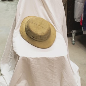 May include: A tan safari hat with a wide brim sits on a white draped surface. The hat has a structured crown and a band around the base. The hat is the focal point, suggesting outdoor or travel-themed accessories.