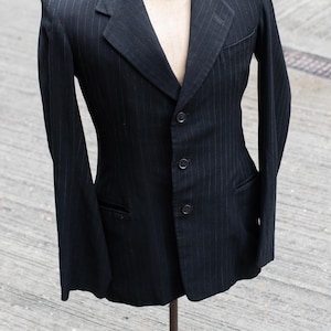 Pode incluir: Um blazer preto com um padrão subtil de risca de giz branca, lapela entalhada e fecho frontal de três botões. A peça é exibida num torso de manequim, apresentando mangas compridas, um bolso no peito e dois bolsos inferiores com aba.