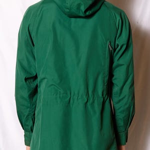Op de afbeelding: Een groene capuchonjas met een ritssluiting en een trekkoord in de taille.