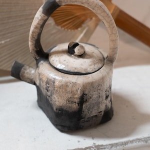 Könnte beinhalten: Eine Keramik-Teekanne mit rundem Henkel und Deckel. Die Teekanne ist überwiegend cremefarben mit schwarzen Akzenten an der Basis, am Henkel und am Deckel. Der Ausguss ist kurz und stämmig. Der Hintergrund umfasst einen Fächer und Holzelemente.