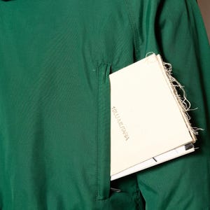 Op de afbeelding: Een donkergroen jack met een ritszak aan de linkerkant. Een wit papiertje met de tekst "1800.1.MILITARIA" is gedeeltelijk zichtbaar in de zak.