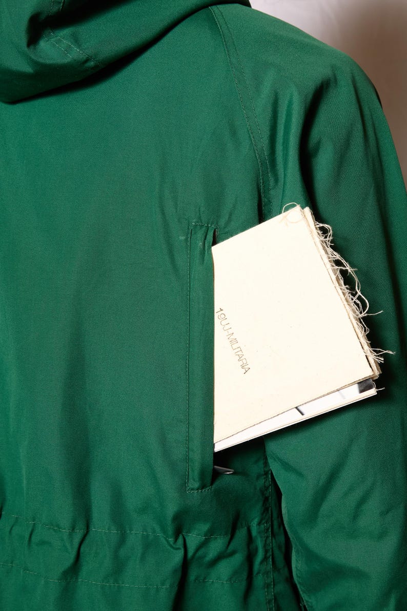 Op de afbeelding: Een donkergroen jack met een ritszak aan de linkerkant. Een wit papiertje met de tekst "1800.1.MILITARIA" is gedeeltelijk zichtbaar in de zak.