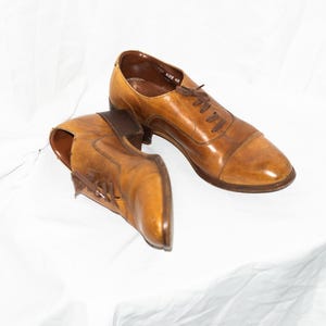 May include: A pair of brown leather dress shoes with laces. The shoes have a cap toe design and are set against a white background. The size is marked as 42.