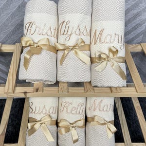 May include: Six rolled, cream-colored towels with diamond patterns are displayed on a wooden rack. Each towel is embroidered with a name in gold thread and tied with a gold satin ribbon. The names are Kristy, Alyssa, Mary, Susan, Kelli, and Mar.