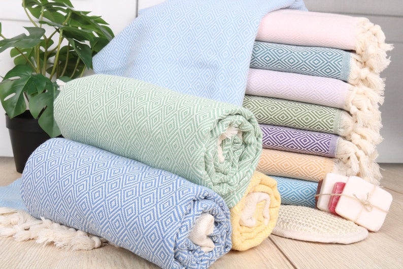 May include: A collection of colorful, rolled and stacked diamond-patterned towels. The towels are in shades of blue, green, yellow, pink, and purple. Some towels are rolled, while others are neatly stacked. The towels have fringed edges.