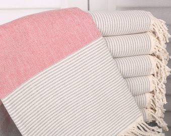 May include: A stack of folded Turkish towels. The towels feature a color block design with a red section, a cream section with gray stripes, and a fringed edge. The towels are made of a woven fabric.