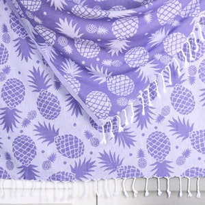 Puede incluir: Una toalla de playa de color morado con un patrón de piña blanca y flecos blancos.