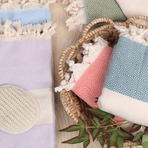 May include: A collection of colorful Turkish towels with diamond patterns and fringed edges. The towels are in shades of blue, lavender, coral, and green, some folded and others in a woven basket. A natural loofah sponge is also present.