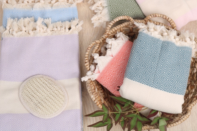 May include: A collection of colorful Turkish towels with diamond patterns and fringed edges. The towels are in shades of blue, lavender, coral, and green, some folded and others in a woven basket. A natural loofah sponge is also present.