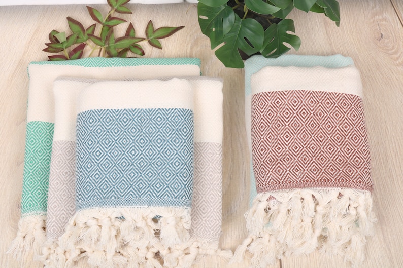 May include: Several folded Turkish towels in shades of blue, green, and cream. The towels feature a diamond pattern and fringe trim. The towels are made of soft, absorbent material, perfect for use in a bathroom or at the beach.