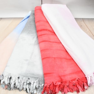 May include: Four colourful fringed towels are fanned out on a light wood surface. The towels are in shades of pink, blue, grey, red, and white. Each towel has a fringed edge.