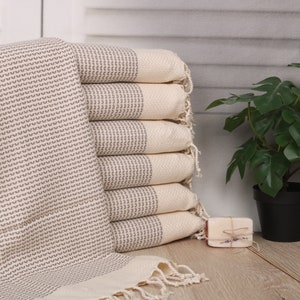 May include: A stack of six beige and gray patterned towels with fringed edges. The towels are neatly arranged, with one towel partially unfolded. A small potted plant and a bar of soap are in the background.