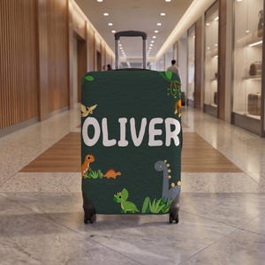Peut inclure: Une housse de valise vert foncé avec le nom OLIVER écrit en grandes lettres blanches. Le motif présente divers dinosaures de dessin animé colorés, dont un dinosaure au long cou gris, un tricératops vert et des dinosaures orange parmi le feuillage.