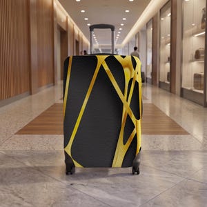 May include: A rolling suitcase featuring a black base colour with a prominent, abstract geometric pattern of intersecting gold lines. The luggage includes a retractable silver handle and four black wheels, standing upright with a smooth, matte finish.