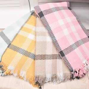 May include: Four colorful, fringed blankets are fanned out on a light wood surface. The blankets are pastel shades of mint green, yellow, beige, and pink. Each blanket has a plaid pattern with a zig-zag border.