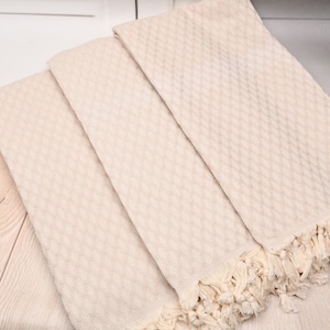 May include: Three rectangular off-white towels featuring a textured diamond weave pattern across the surface. Each towel is finished with a row of knotted fringe along the bottom edge, creating a consistent look across the set of textile items.