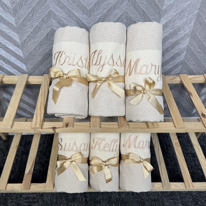 May include: Six rolled, cream-colored towels with diamond patterns are displayed on a wooden rack. Each towel is embroidered with a name in gold thread and tied with a gold satin ribbon. The names are Kristy, Alyssa, Mary, Susan, Kelli, and Mar.