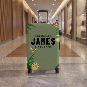 May include: A sage green suitcase cover featuring a dinosaur theme with palm leaves, a hatching egg, and a flying pterodactyl. The text reads HELLO, MY NAME IS JAMES and JAMES DAVIS in bold black lettering centered on the front of the luggage.