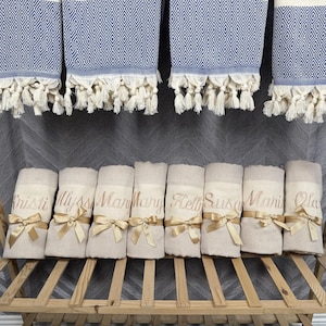 May include: A display of personalized beige and blue diamond-patterned towels. The towels are rolled and tied with gold ribbons, each embroidered with a name. Above, matching blue and white patterned towels hang with fringe.