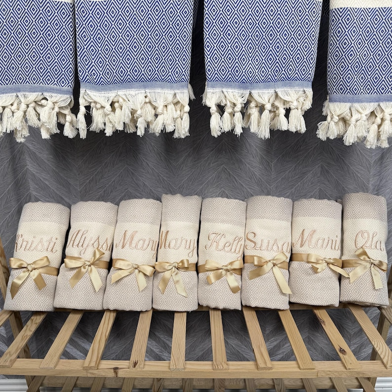 May include: A display of personalized beige and blue diamond-patterned towels. The towels are rolled and tied with gold ribbons, each embroidered with a name. Above, matching blue and white patterned towels hang with fringe.