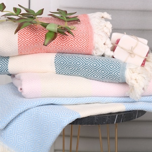 May include: A stack of colorful Turkish towels with diamond patterns and fringe. The towels are in shades of coral, teal, pink, and light blue. A sprig of greenery and a small bar of soap are placed on top of the towels.