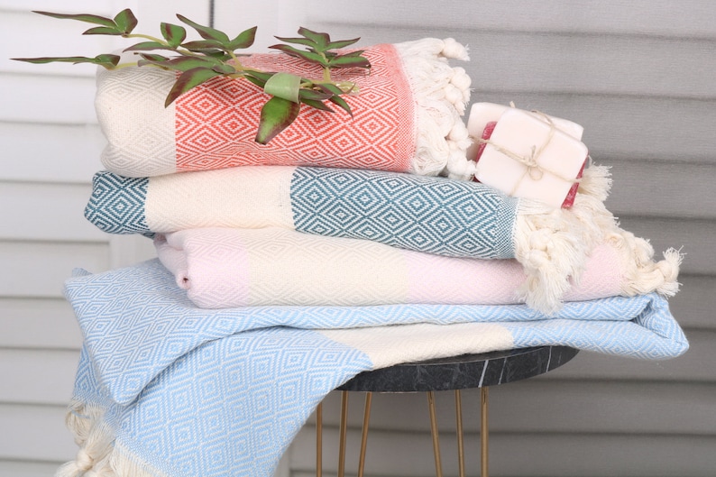 May include: A stack of colorful Turkish towels with diamond patterns and fringe. The towels are in shades of coral, teal, pink, and light blue. A sprig of greenery and a small bar of soap are placed on top of the towels.