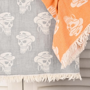 May include: Two decorative towels, one grey and one orange, feature a repeating skull design wearing a cowboy hat and earrings. The towels have a fringed edge. The grey towel is hanging, and the orange towel is folded.