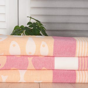 May include: Three folded beach towels stacked on top of each other. The towels have a pattern of white shapes on a peach background, with a pink stripe and a striped section. The towels have fringed edges.