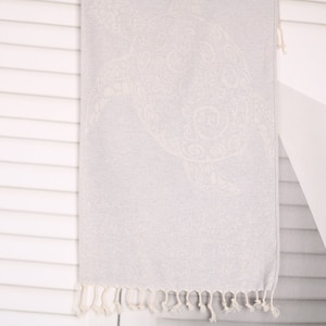 May include: A light gray beach towel with a detailed turtle design. The towel has a fringe along the bottom edge and is hanging against a white slatted wall. The turtle design is a lighter shade of gray.