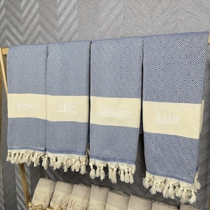 May include: Four blue and white diamond patterned beach towels with cream-colored bands embroidered with the names "Michelle", "Ashley", "Julianne", and "Katie". Each towel has a fringe along the bottom edge.