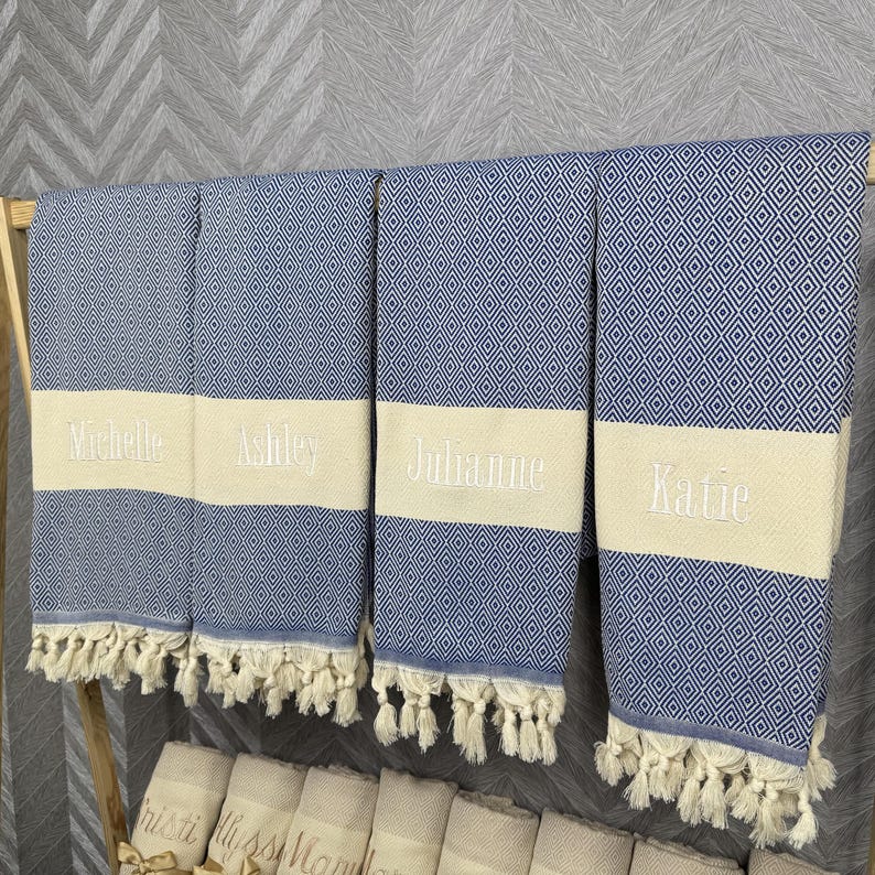 May include: Four blue and white diamond patterned beach towels with cream-colored bands embroidered with the names "Michelle", "Ashley", "Julianne", and "Katie". Each towel has a fringe along the bottom edge.