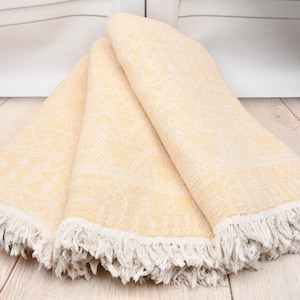 Könnte beinhalten: Ein gefaltetes hellgelbes Textil mit einem gewebten geometrischen Muster und weißen Fransenrändern. Der Stoff zeigt ein sich wiederholendes Design aus Dreiecken und linearen Motiven auf der Oberfläche, abgeschlossen mit langen, geknoteten weißen Quasten.