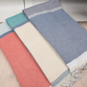 May include: Three folded beach towels in coral, teal, and blue. Each towel has a contrasting border and fringe detail. The towels are made of a textured fabric and are arranged on a light wood surface.