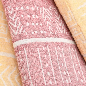 Peut inclure: Gros plan sur trois serviettes en coton pliées. L'une est rouge clair avec des motifs géométriques blancs, une autre est jaune avec des motifs géométriques blancs, et la troisième est jaune clair avec des motifs géométriques blancs. Les serviettes ont un aspect texturé.