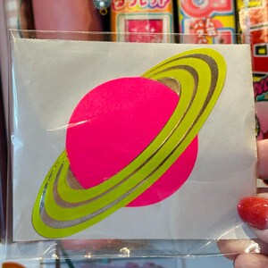 May include: A neon pink planet sticker with a bright green and silver ring design. The sticker is on a white background and is in a clear plastic sleeve. The sticker is a fun, colorful design.
