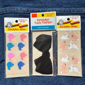 May include: Three Sandylion sticker packs in clear plastic sleeves. The left pack features pink and blue ice cream cone stickers. The centre pack contains a large black fuzzy bow sticker. The right pack displays white and tan bunny rabbit stickers.
