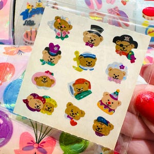 May include: A sheet of vintage stickers featuring various teddy bears in different outfits. The bears are wearing hats, sailor outfits, and pirate costumes. The stickers are in a clear plastic sleeve, with other sticker sheets visible in the background.