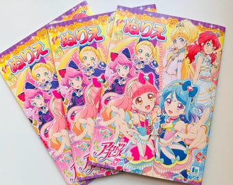 May include: Four colourful colouring books featuring anime-style girls with pink, blue, and yellow hair. The covers have Japanese text and decorative borders in pink and purple. The books are stacked on a white surface.