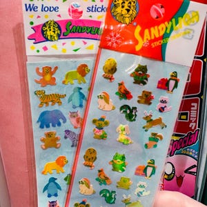 Könnte beinhalten: Zwei Stickerbögen mit bunten Tierdesigns. Ein Bogen hat ein Löwenlogo und den Text "We love stick SANDYLI". Der andere Bogen hat ein Löwenlogo und den Text "SANDYLIO STICKER". Die Sticker zeigen Löwen, Tiger, Pinguine und mehr.