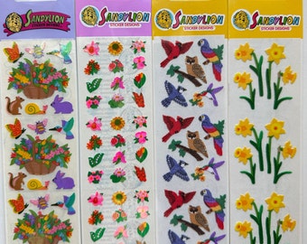 May include: Four sheets of Sandy Lion sticker designs. The sheets feature various designs, including butterflies, flowers, birds, and animals. The stickers have a shiny, pearlescent finish and a fuzzy texture. The top of each sheet has the Sandy Lion logo and the words "Mother of Pearl" or "Fuzzy Velours".