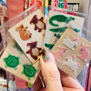 May include: A collection of felt animal stickers, including koalas, turtles, alligators, and rabbits. The stickers are in various colors, such as green, brown, orange, and pink. Each sticker is on a small, square piece of cardboard.