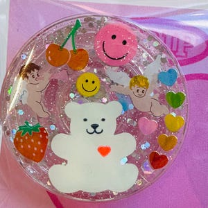 May include: A clear, round resin charm with a white teddy bear, cherubs, cherries, a strawberry, hearts, and smiley faces. The charm is filled with glitter and has a pink background.