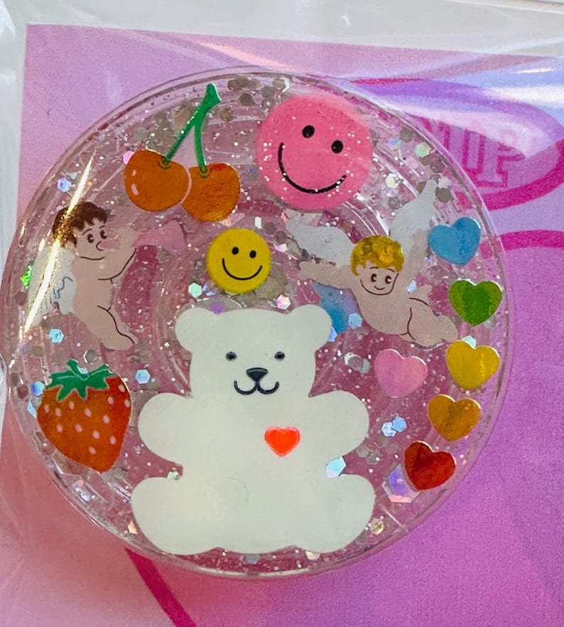 May include: A clear, round resin charm featuring a white teddy bear, cherubs, cherries, a strawberry, hearts, and smiley faces. The charm is filled with glitter and has a pink background.